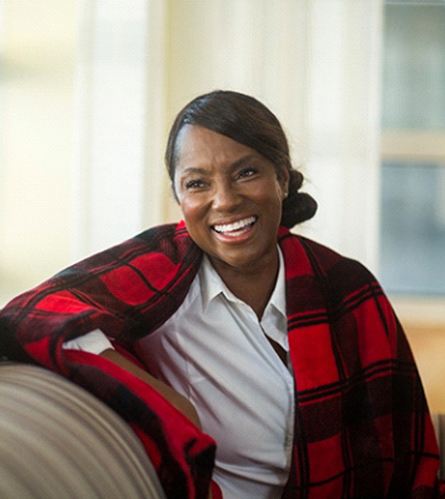 A middle-aged woman sitting on a couch with a red and black shawl over her shoulders, smiling and pleased with her new implant dentures in Ellicott City