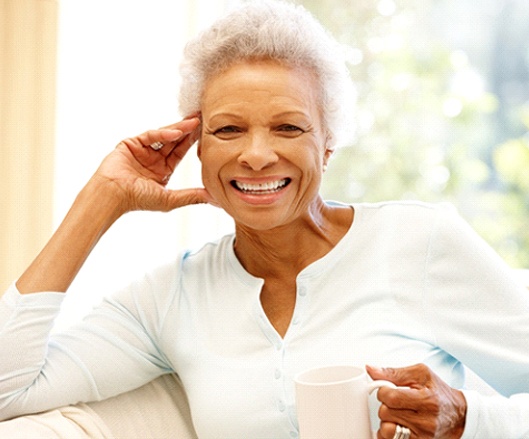 An older woman sitting on a couch and holding a cup in her hand while smiling and showing off her new teeth