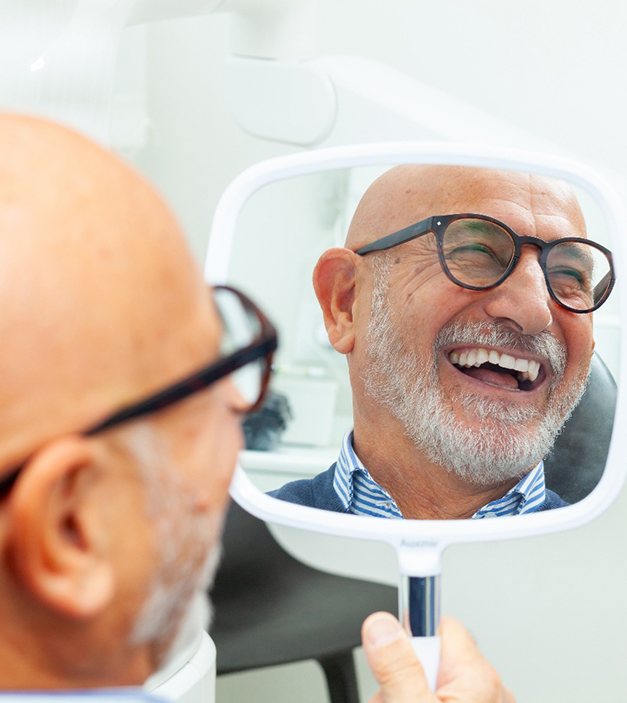 a smiling man looking in a handheld mirror in a dental office